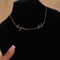 A delicate gold script necklace spelling 'kiyomi' and 'kiki' displayed on a black velvet jewelry bust, with a hand holding the side of the bust. The background shows a light wood floor.